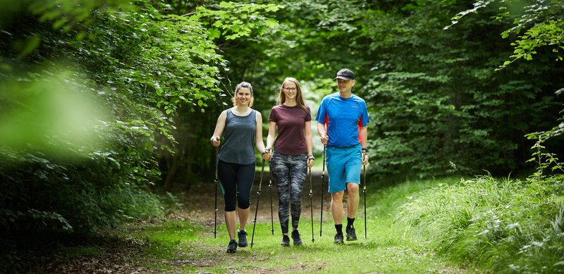 Zwei Frauen und ein Mann in Sportkleidung laufen mit Stöcken auf einem Waldweg