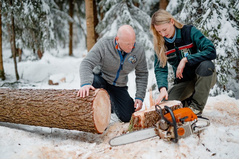 Försterin und Waldbesitzer knien im verschneiten Wald vor einem Wurzelstock. Im Vordergrund ein gefällter Baum und eine Motorsäge.
