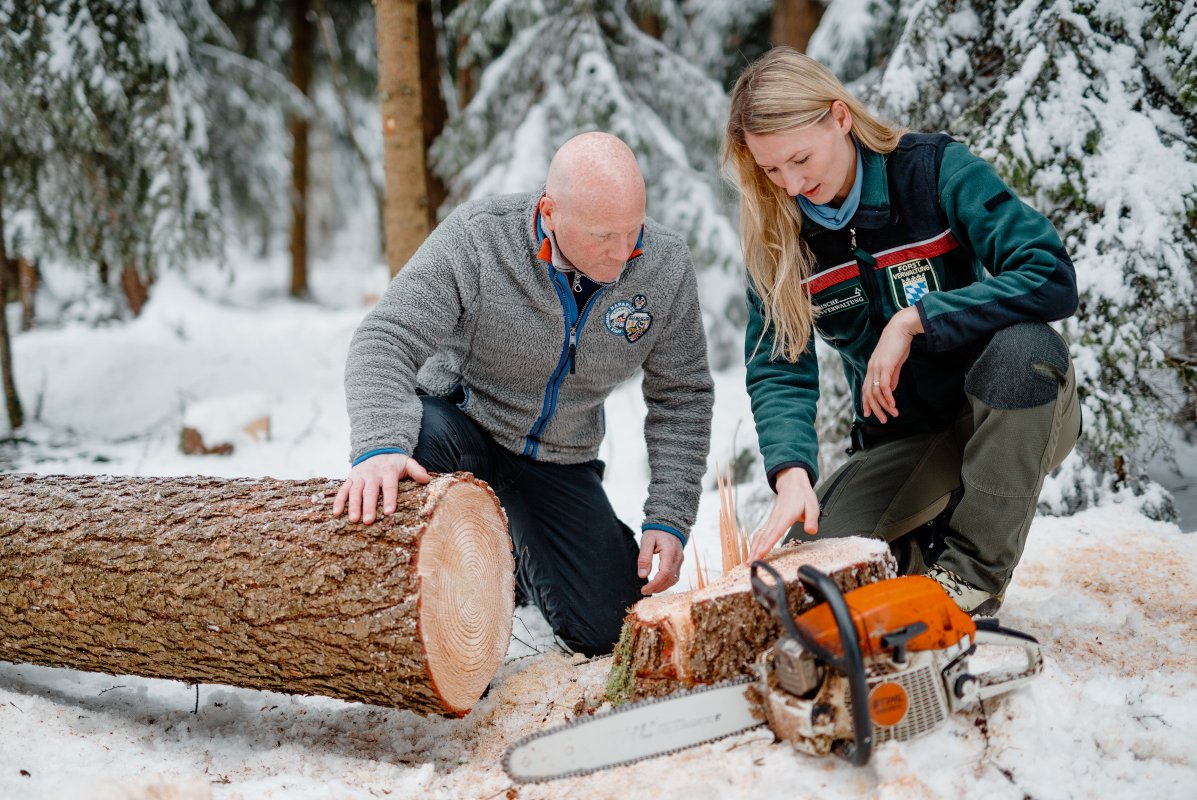 Försterin und Waldbesitzer knien im verschneiten Wald vor einem Wurzelstock. Im Vordergrund ein gefällter Baum und eine Motorsäge.