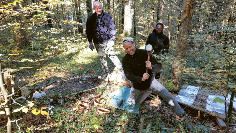 Drei Männer im Wald mit einer Versuchsanordnung, um Kohlenstoff zu messen. Einer von ihnen hebt spielerisch einen Hammer in die Kamera 