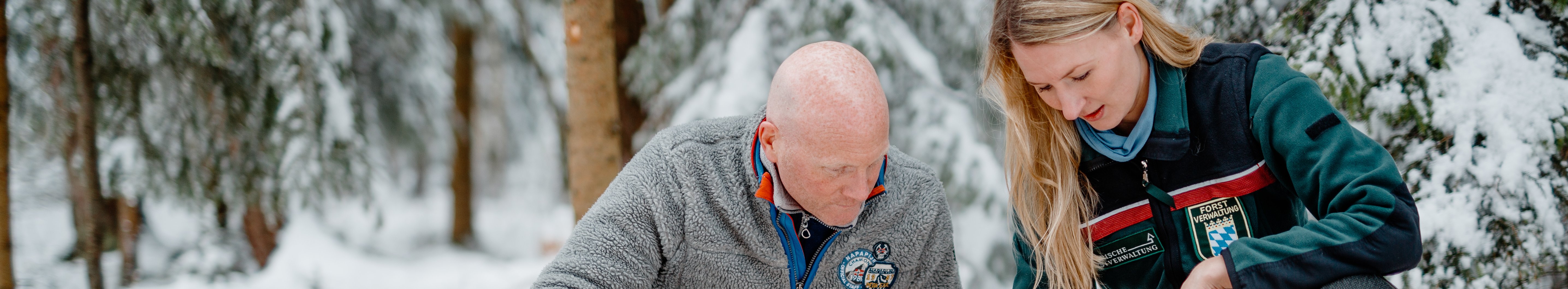 Försterin und Waldbesitzer knien im verschneiten Wald vor einem Wurzelstock. Im Vordergrund ein gefällter Baum und eine Motorsäge.