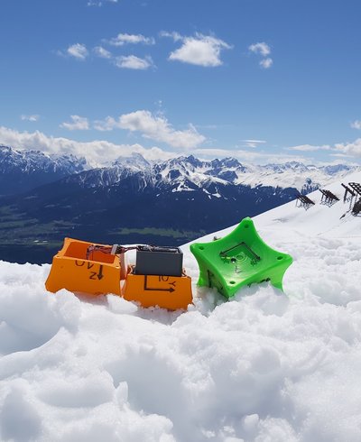Orangene und grüne Würfel zur Lawinenforschung liegen auf einem schneebedeckten Berg, mit Blick ins Tal
