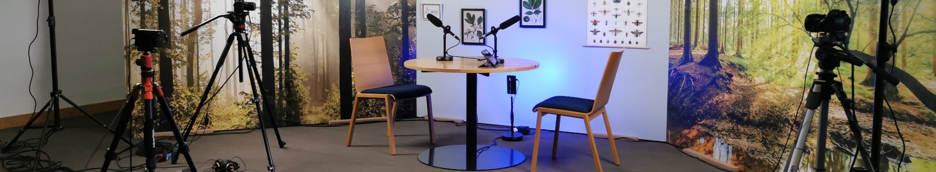 Aufnahmeset des Videopodcast mit zwei Stühlen, einem Tisch und einer Wald-Fototapete im Hintergrund