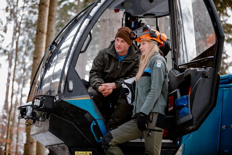 Försterin steht neben einer Harvester-Kanzel und gibt dem Fahrer Anweisungen